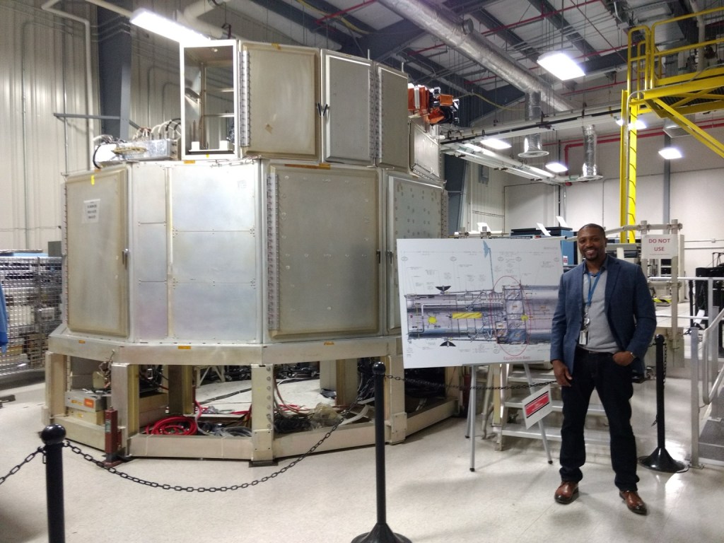 Ben Teasdel III standing beside spacecraft hardware and a technical diagram in an engineering facility